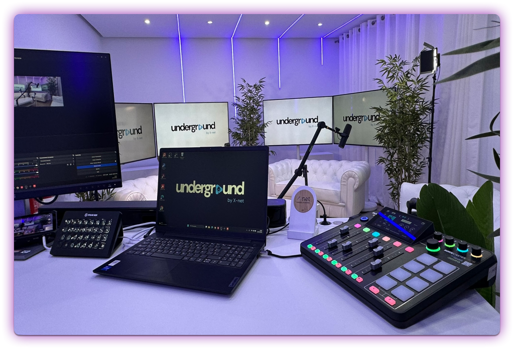 Vista de un estudio de podcasting equipado con una consola RØDECaster Pro II, laptop Lenovo, Stream Deck, micrófonos profesionales y pantallas de fondo con el logo Underground by X-net.